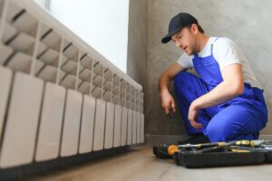 A man in overalls kneels beside a radiator, inspecting it with focused attention in a well-lit room.